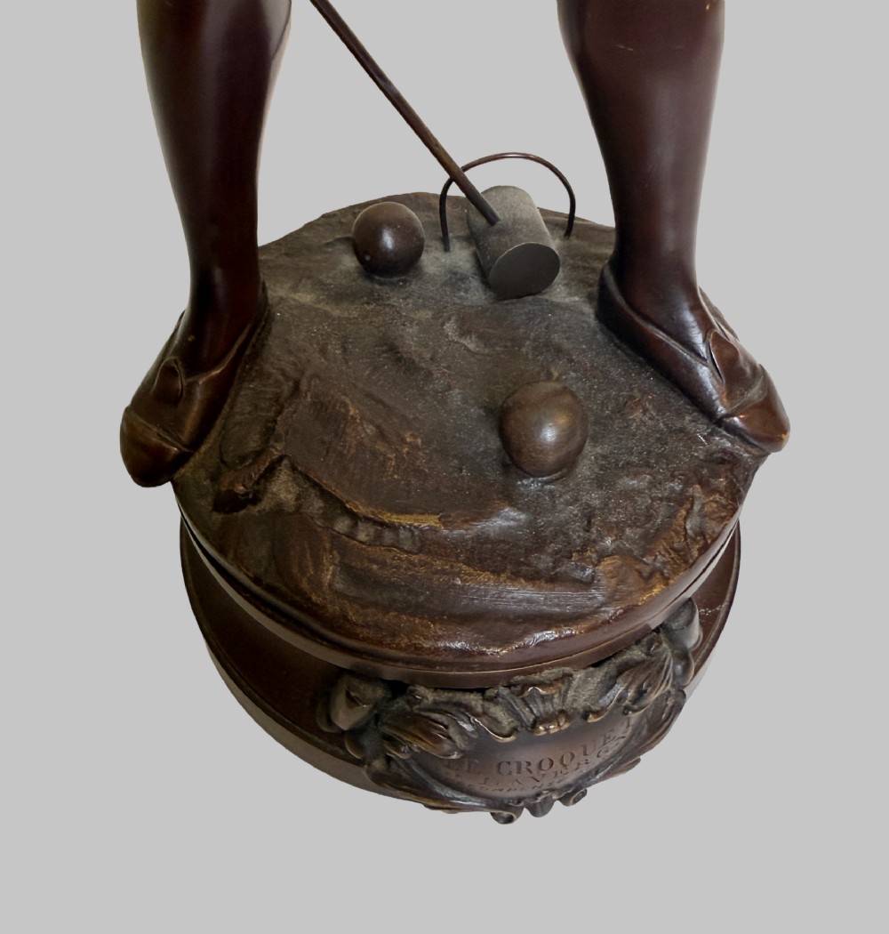 A dark patinated bronze statue of a young man smoking a cigarette while holding a croquet mallet on a round base with croquet balls. View VIII