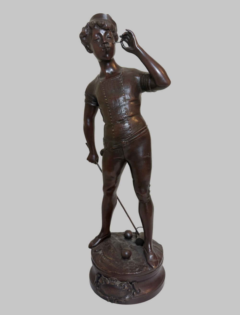 A dark patinated bronze statue of a young man smoking a cigarette while holding a croquet mallet on a round base with croquet balls. View IX