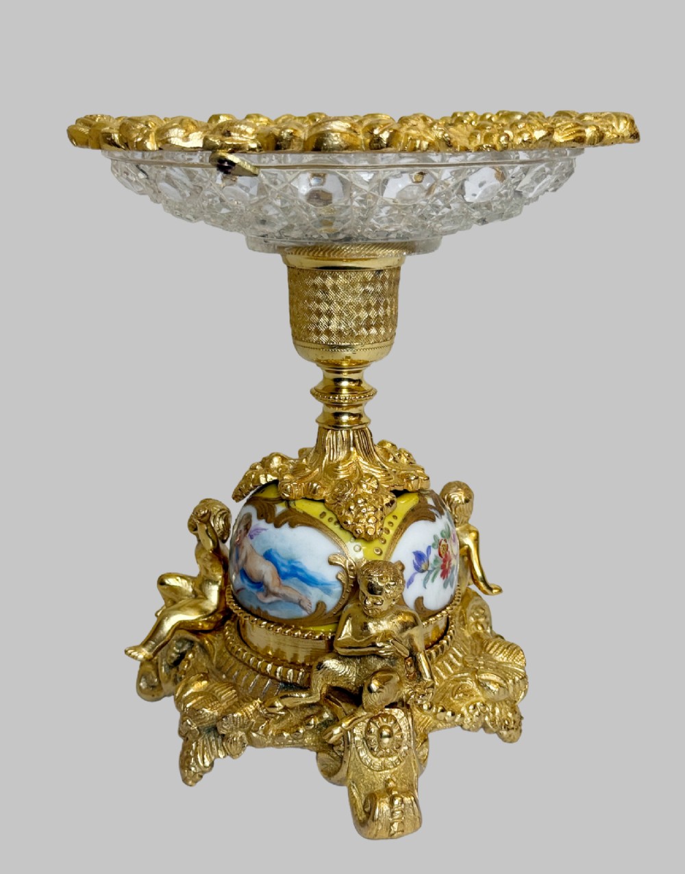 An ornate gilt bronze centrepiece dish featuring a cut crystal bowl, hand-painted porcelain cherub panels, and three seated bronze putti figures on a scrolled base. View X