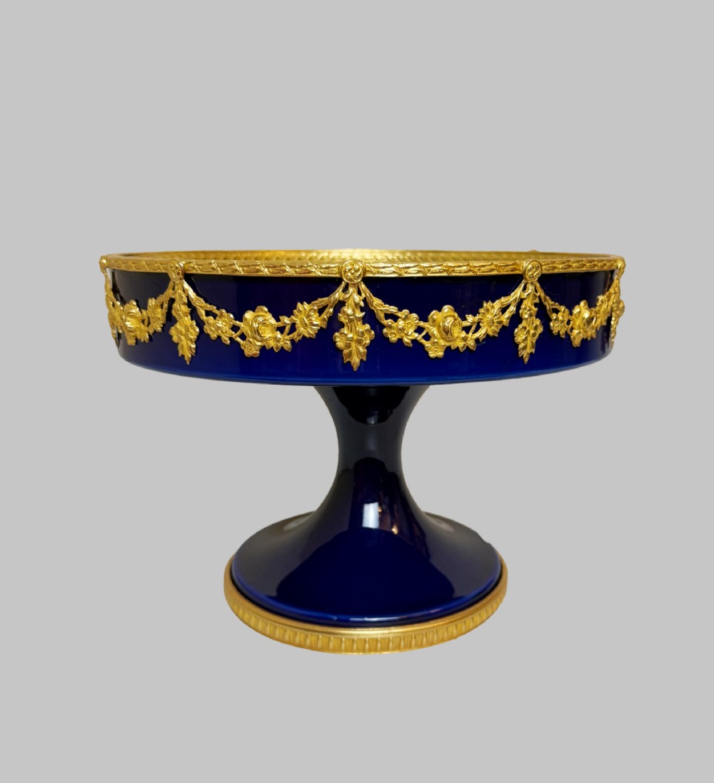 A deep cobalt blue porcelain bowl raised on a pedestal base with ornate gilt bronze floral swags and a chased rim. View II