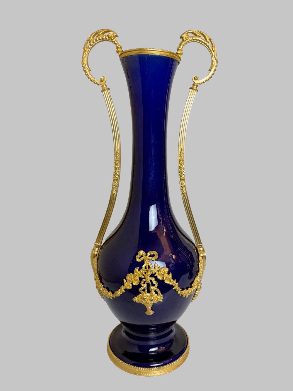 A pair of slender 19th-century cobalt blue porcelain vases featuring high elongated necks, leaf-scroll gilt bronze handles, and gilded floral bow swags around the base. View II