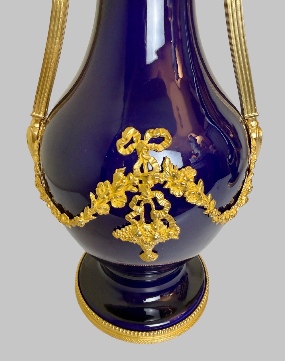 A pair of slender 19th-century cobalt blue porcelain vases featuring high elongated necks, leaf-scroll gilt bronze handles, and gilded floral bow swags around the base. View V