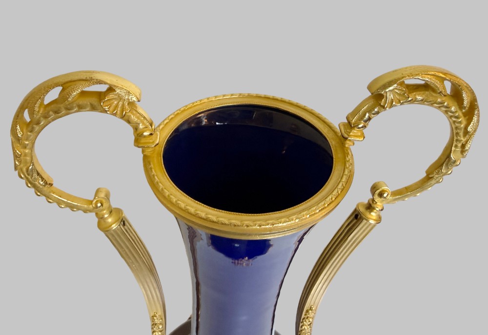 A pair of slender 19th-century cobalt blue porcelain vases featuring high elongated necks, leaf-scroll gilt bronze handles, and gilded floral bow swags around the base. View VI