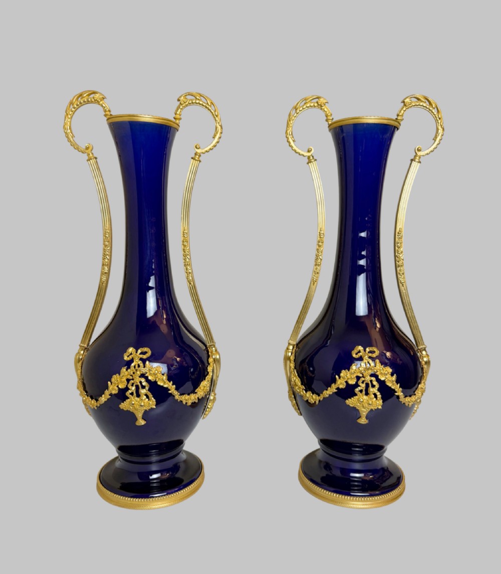 A pair of slender 19th-century cobalt blue porcelain vases featuring high elongated necks, leaf-scroll gilt bronze handles, and gilded floral bow swags around the base. View VII