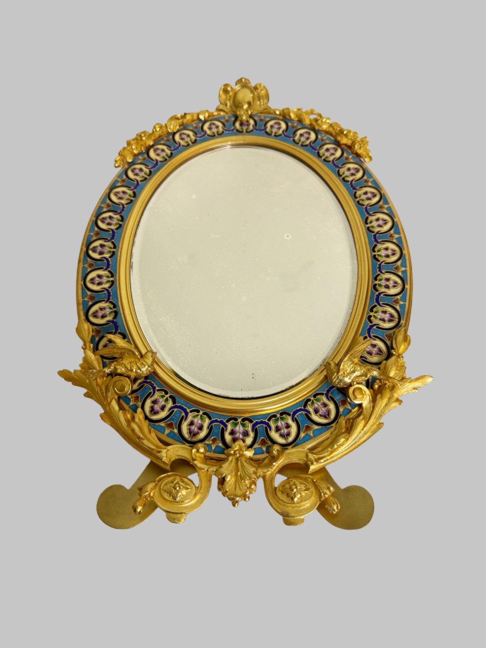 A late 19th-century oval dressing table mirror featuring a gilt bronze frame adorned with blue and purple champleve enamel and bird motifs. View X