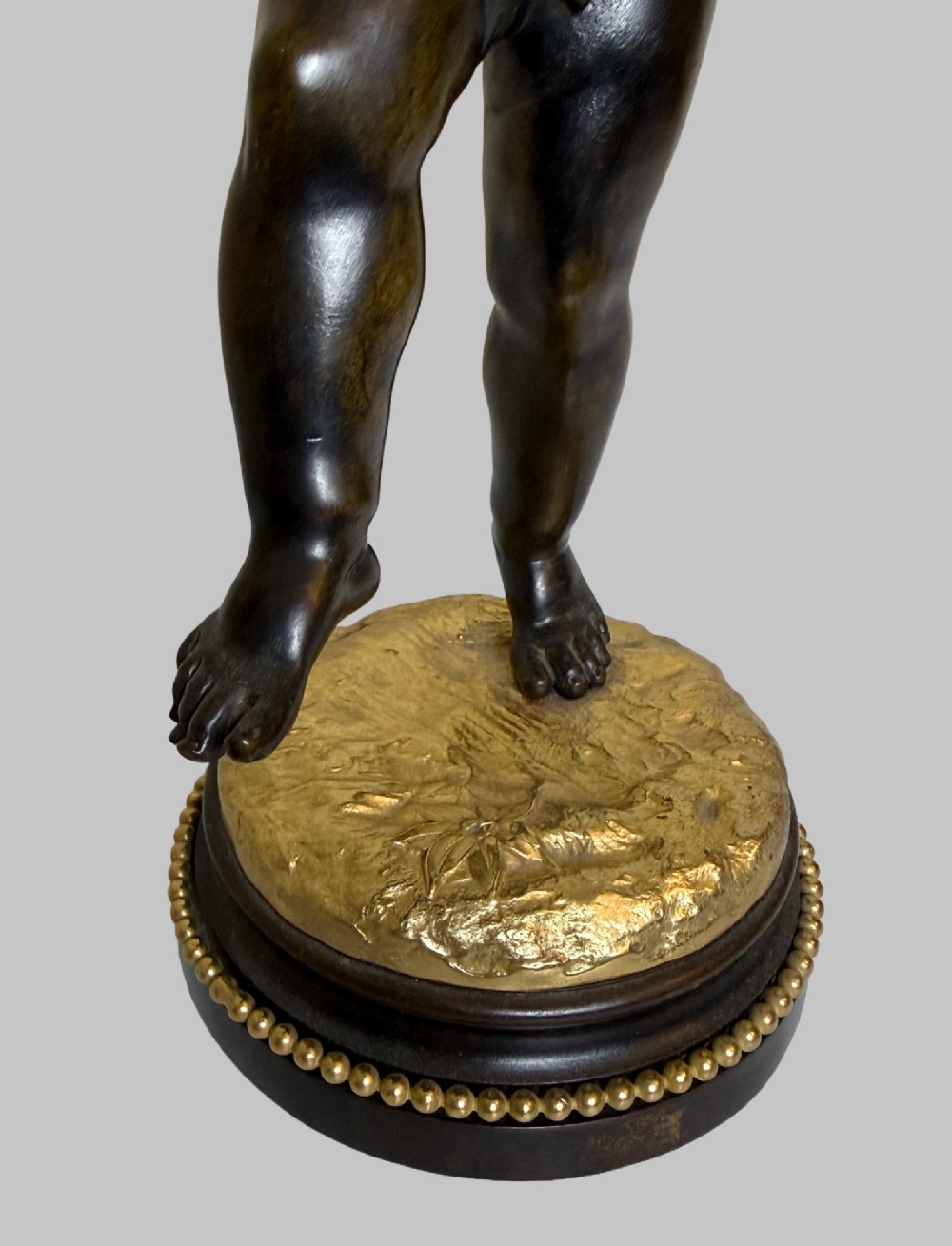 A dark patinated bronze figure of a cherub playing a musical triangle standing on a gilt base mounted on a circular dark wooden plinth. View II