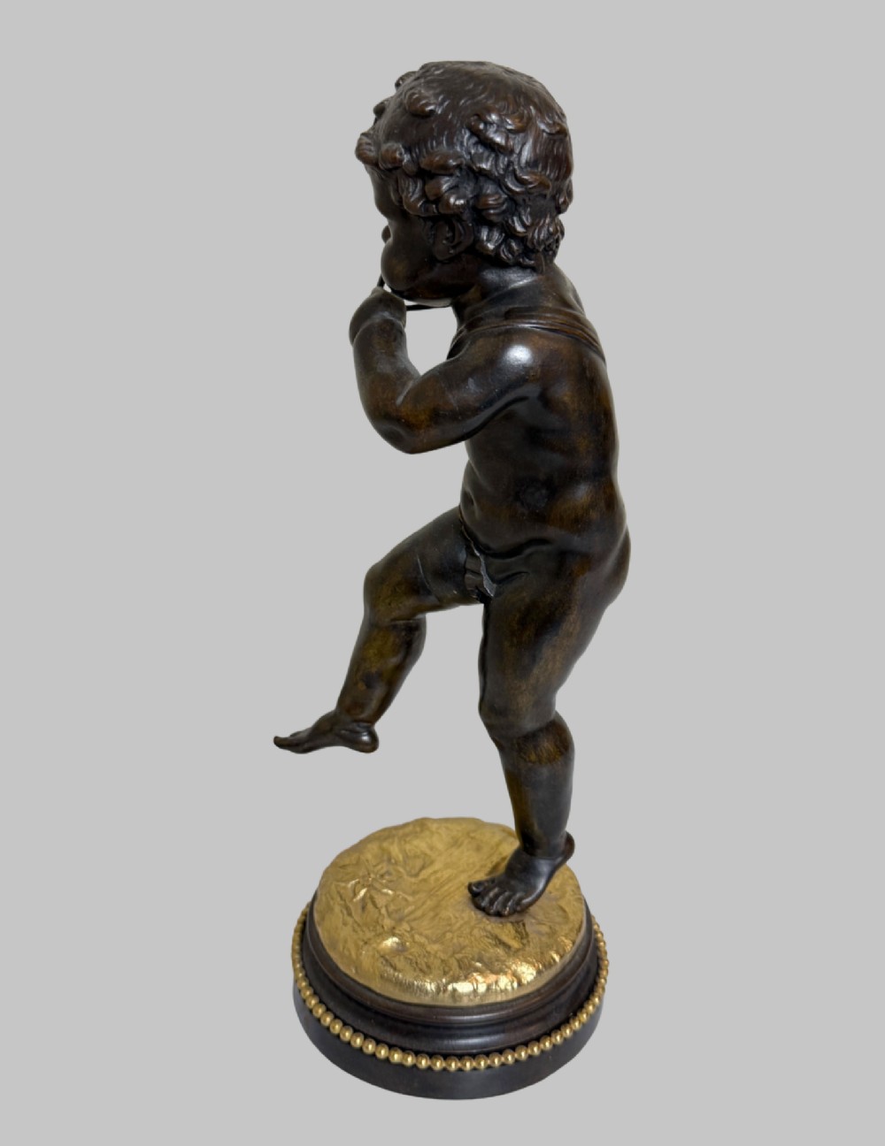 A dark patinated bronze figure of a cherub playing a musical triangle standing on a gilt base mounted on a circular dark wooden plinth. View IV