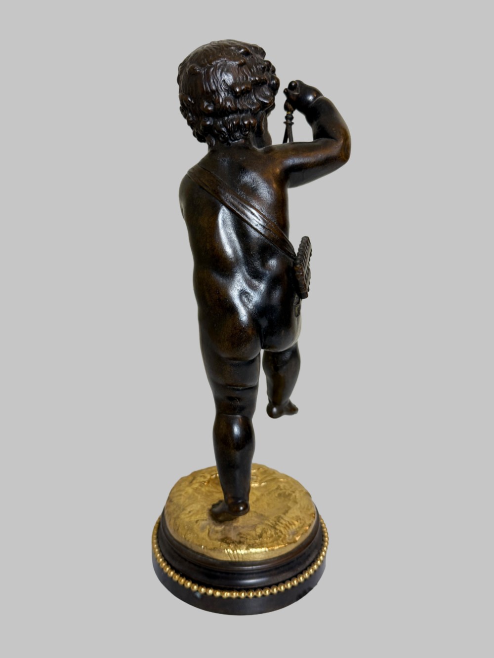 A dark patinated bronze figure of a cherub playing a musical triangle standing on a gilt base mounted on a circular dark wooden plinth. View V