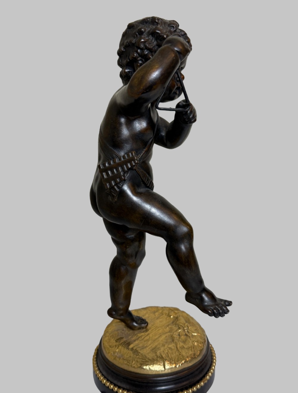 A dark patinated bronze figure of a cherub playing a musical triangle standing on a gilt base mounted on a circular dark wooden plinth. View VI