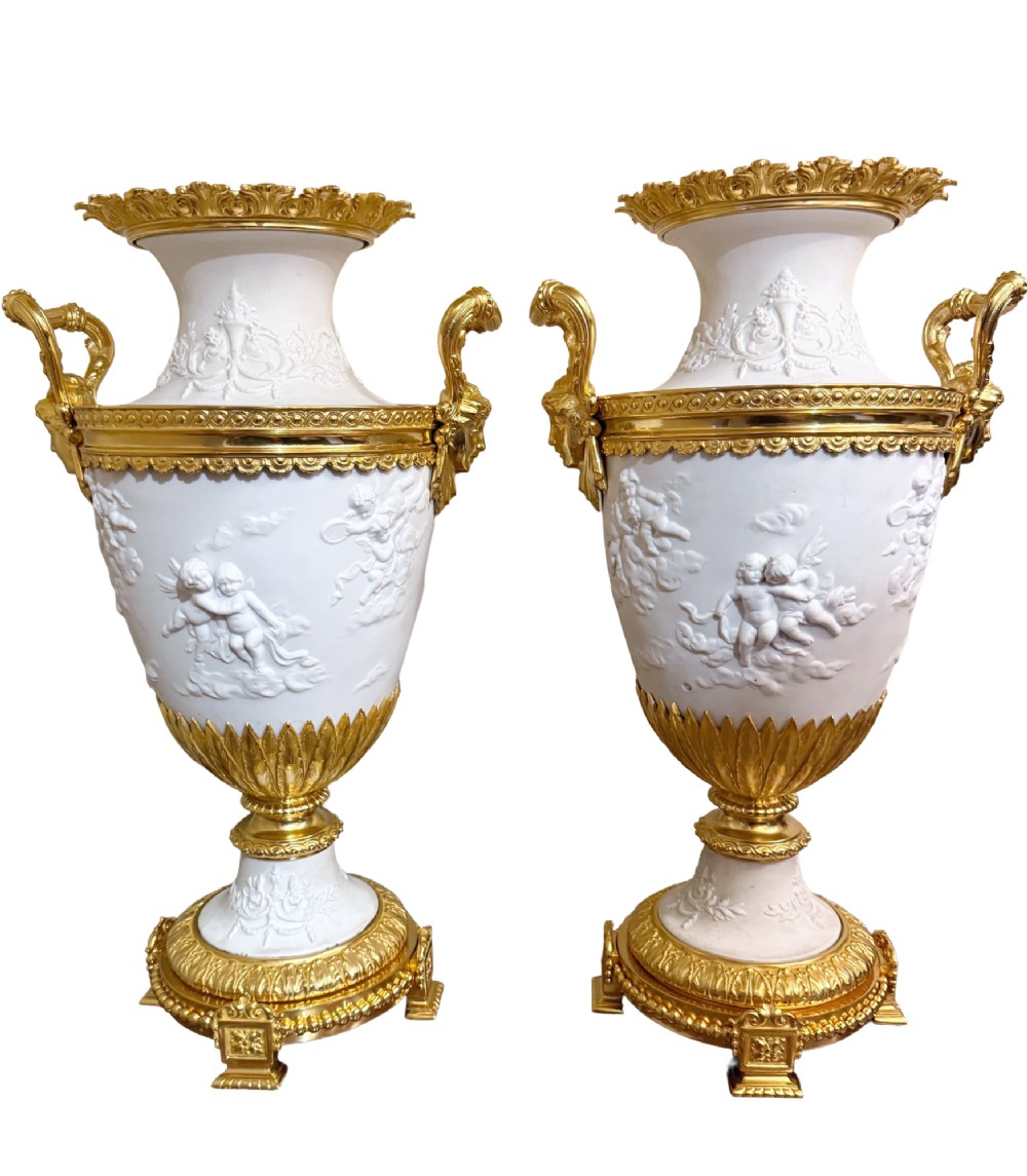 A pair of white porcelain urns adorned with raised relief putti motifs and detailed gilt bronze bases, handles, and rims.