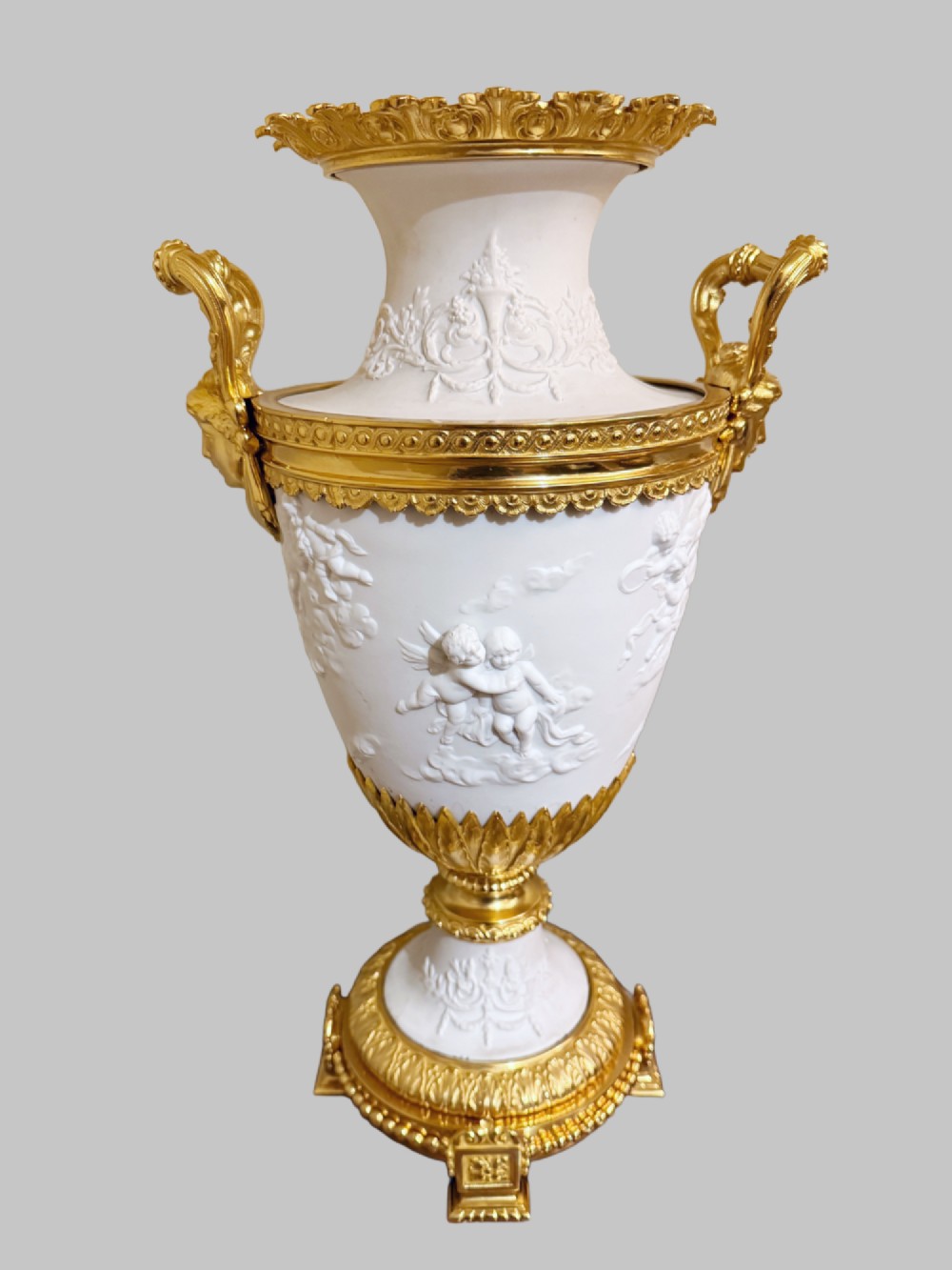 A pair of white porcelain urns adorned with raised relief putti motifs and detailed gilt bronze bases, handles, and rims. View II