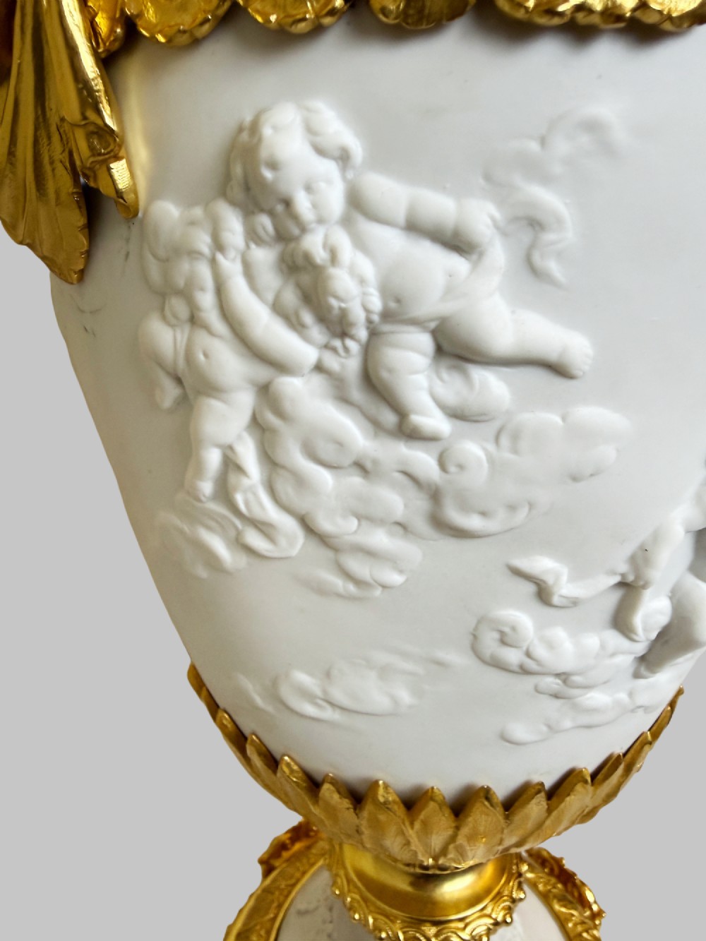 A pair of white porcelain urns adorned with raised relief putti motifs and detailed gilt bronze bases, handles, and rims. View IV