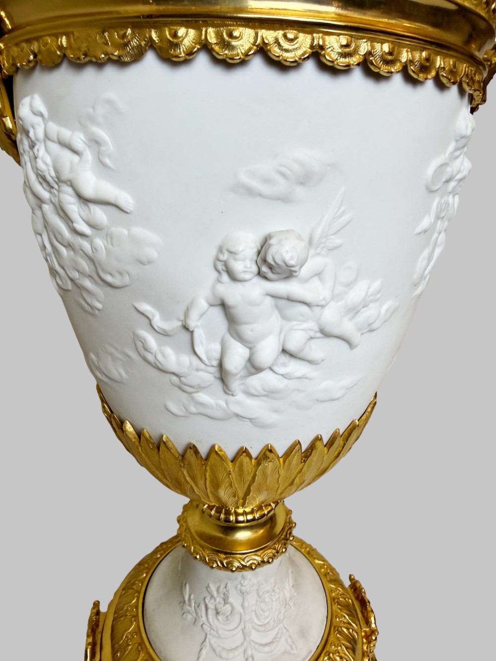 A pair of white porcelain urns adorned with raised relief putti motifs and detailed gilt bronze bases, handles, and rims. View VI