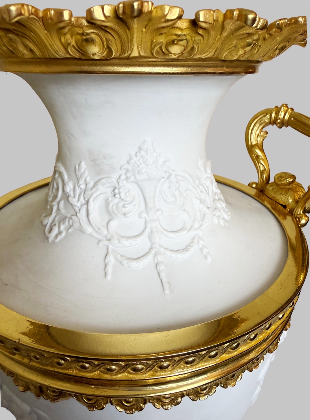 A pair of white porcelain urns adorned with raised relief putti motifs and detailed gilt bronze bases, handles, and rims. View VII