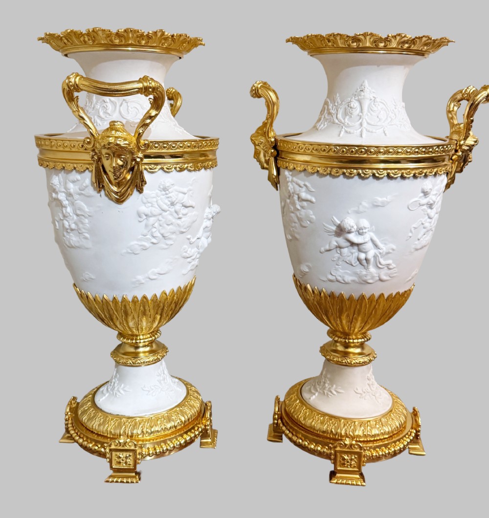 A pair of white porcelain urns adorned with raised relief putti motifs and detailed gilt bronze bases, handles, and rims. View IX