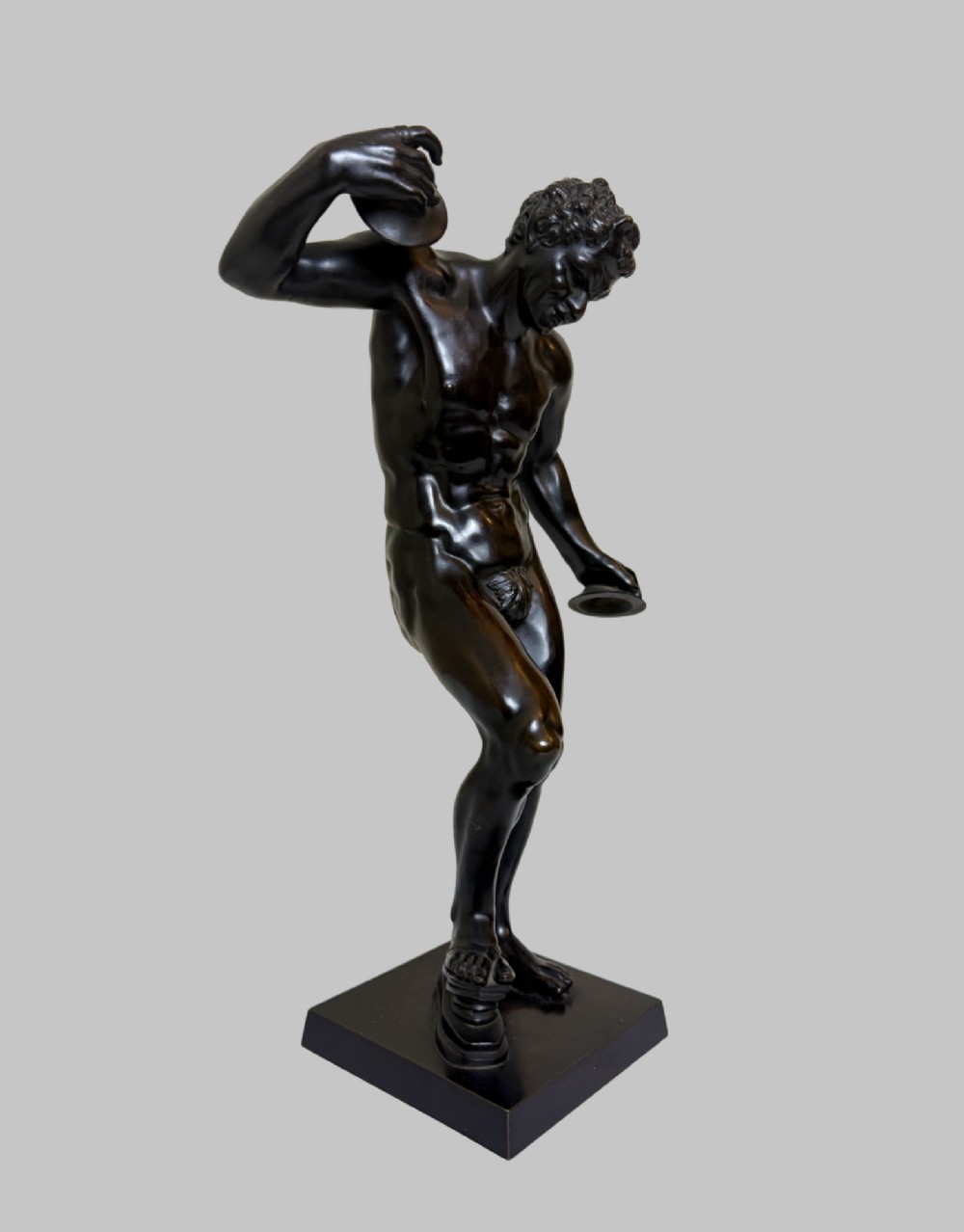 A dark patinated bronze statue depicting a nude classical dancing faun holding cymbals, mounted on a square black base. View II