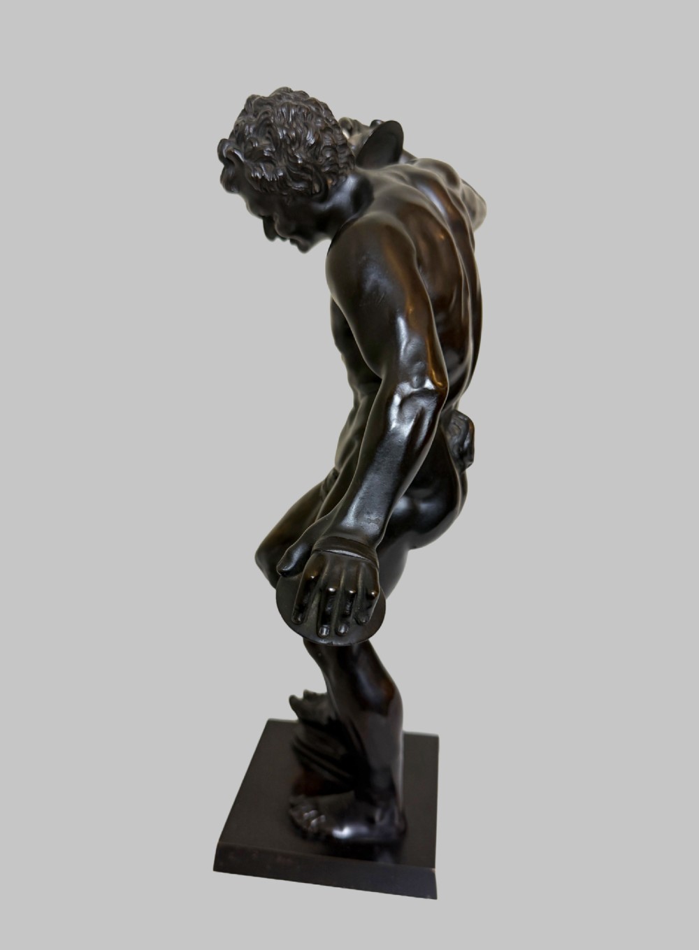 A dark patinated bronze statue depicting a nude classical dancing faun holding cymbals, mounted on a square black base. View IV
