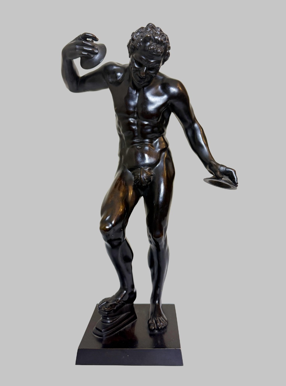 A dark patinated bronze statue depicting a nude classical dancing faun holding cymbals, mounted on a square black base. View X