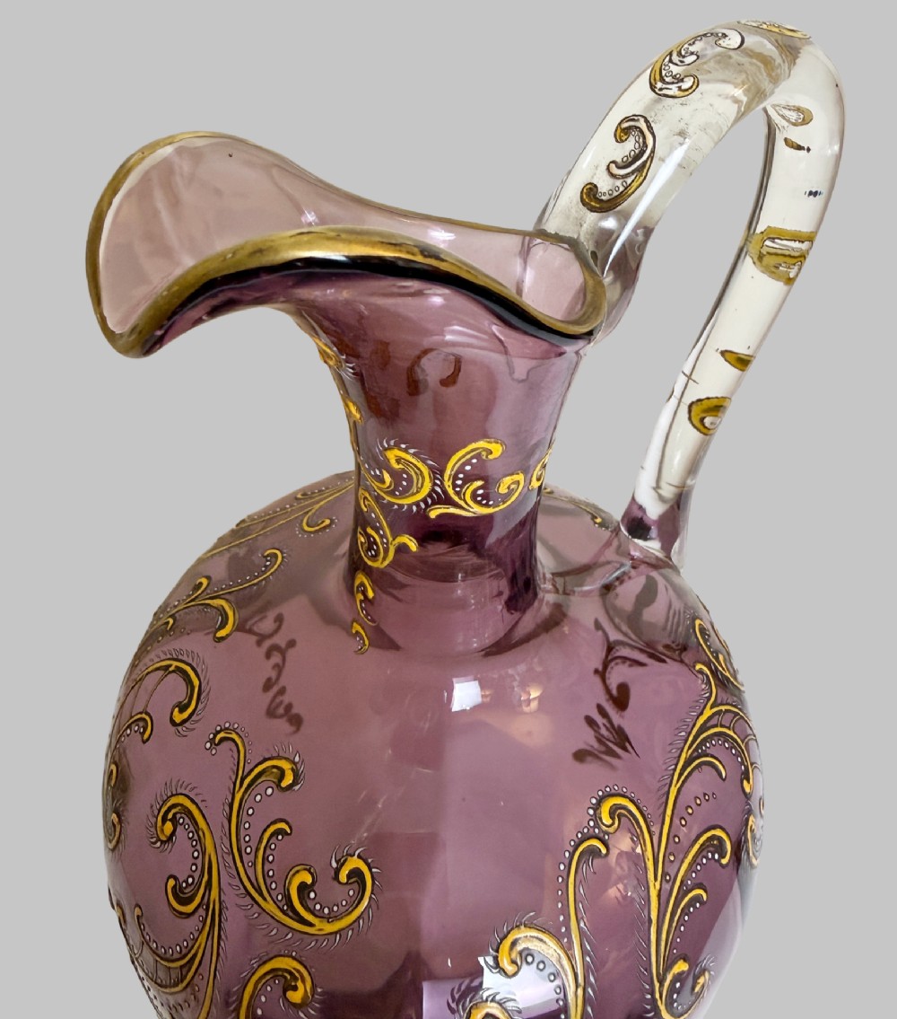 A 19th-century blown violet glass ewer featuring a clear glass handle, slender neck, circular base, and hand-painted gilt scroll patterns. View IV