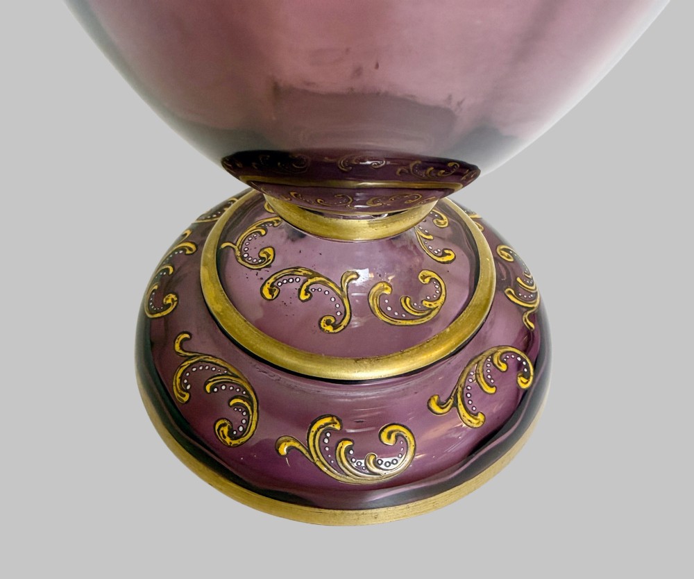 A 19th-century blown violet glass ewer featuring a clear glass handle, slender neck, circular base, and hand-painted gilt scroll patterns. View VI