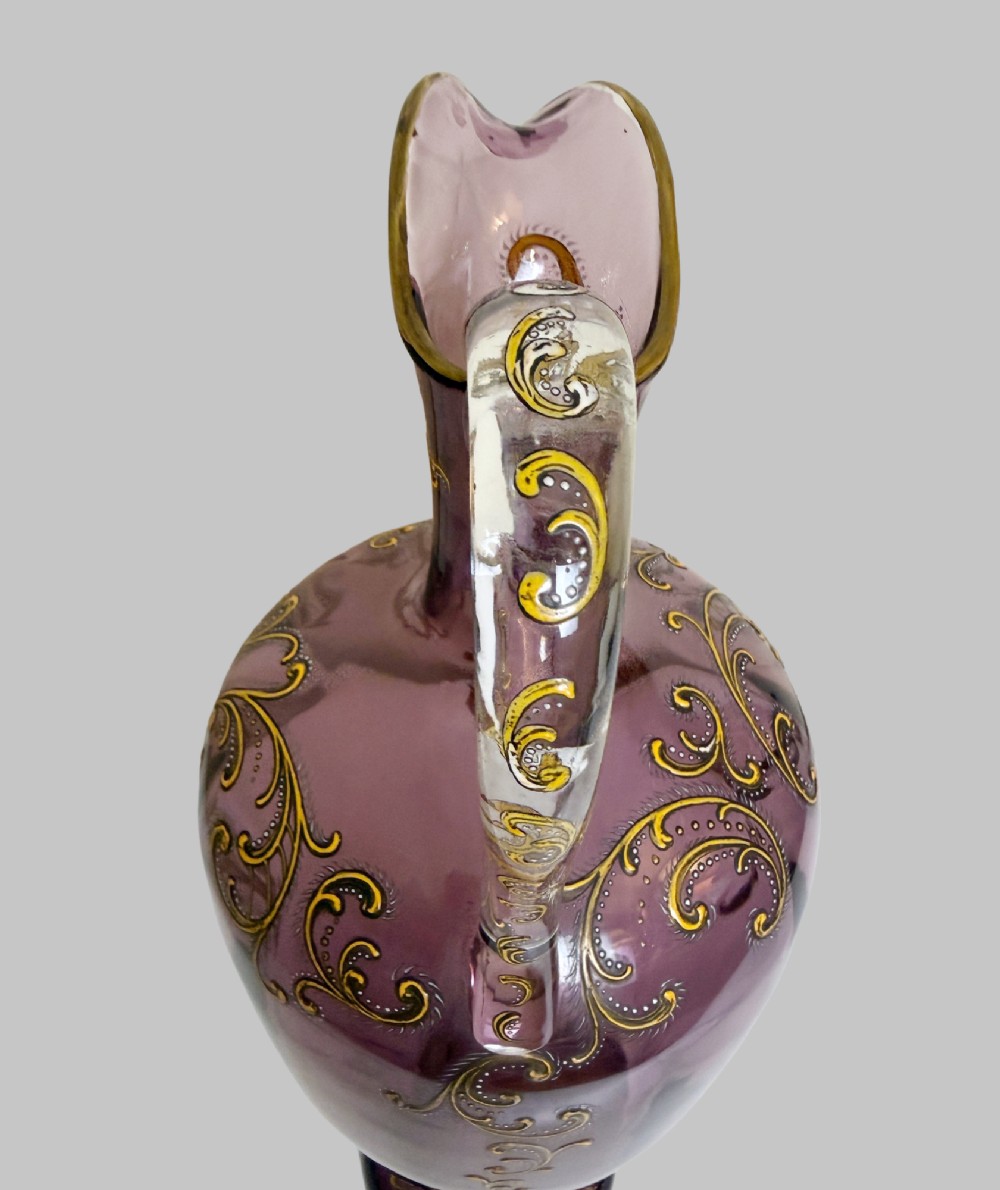 A 19th-century blown violet glass ewer featuring a clear glass handle, slender neck, circular base, and hand-painted gilt scroll patterns. View VII