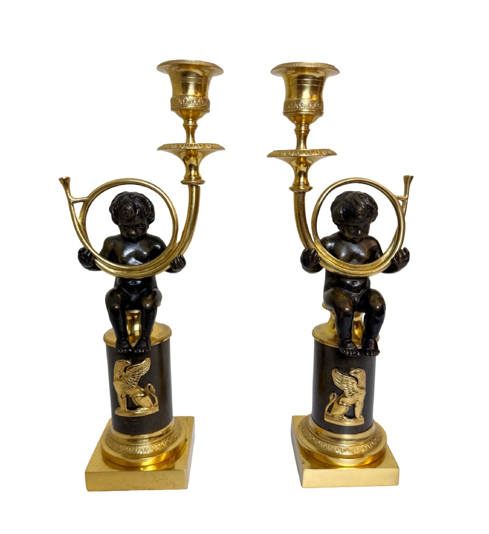 A pair of 19th-century gilt and patinated bronze candlesticks featuring dark cherubs seated on cylindrical pedestals with gilt winged lion motifs, holding gold-toned horn sconces.