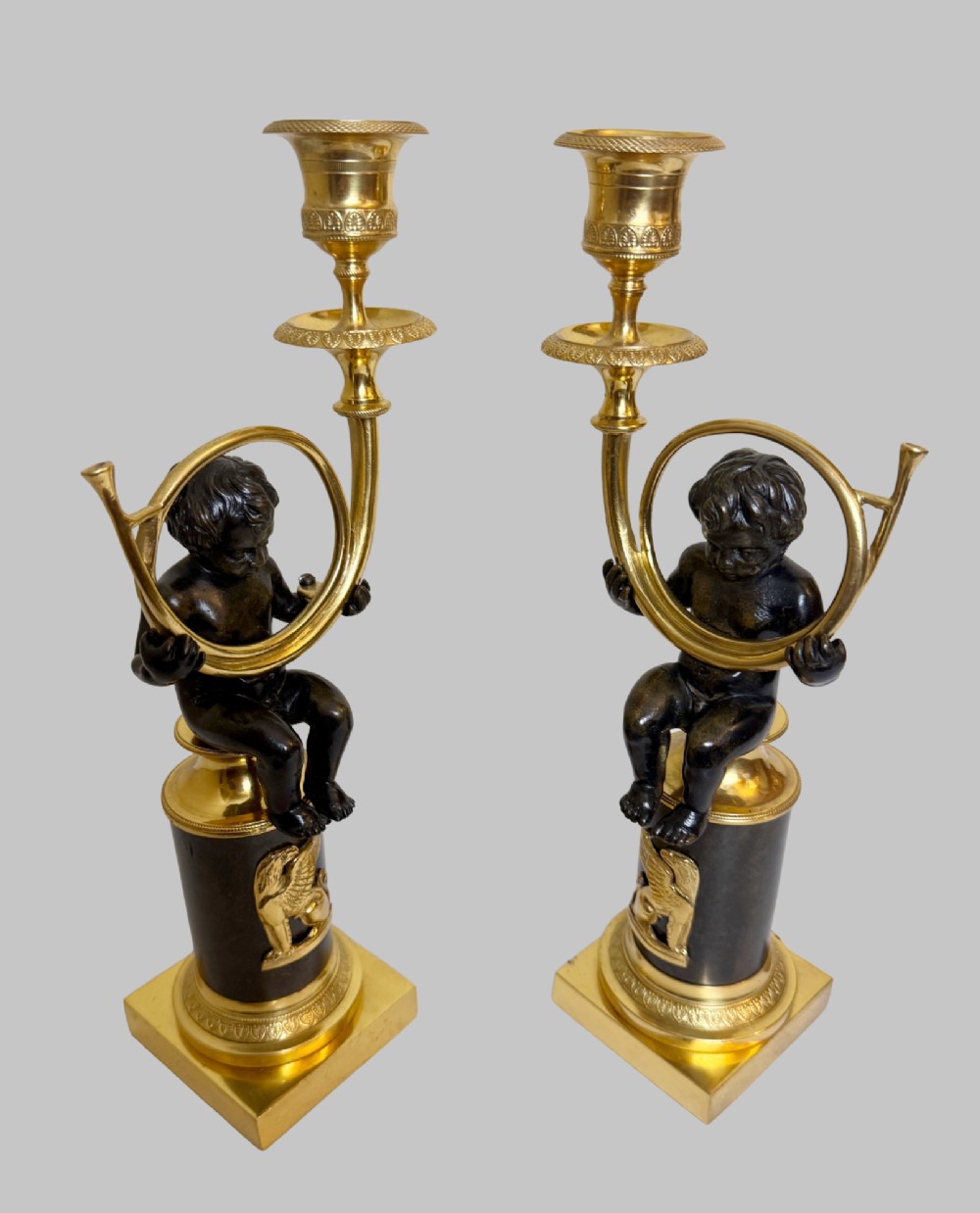 A pair of 19th-century gilt and patinated bronze candlesticks featuring dark cherubs seated on cylindrical pedestals with gilt winged lion motifs, holding gold-toned horn sconces. View II