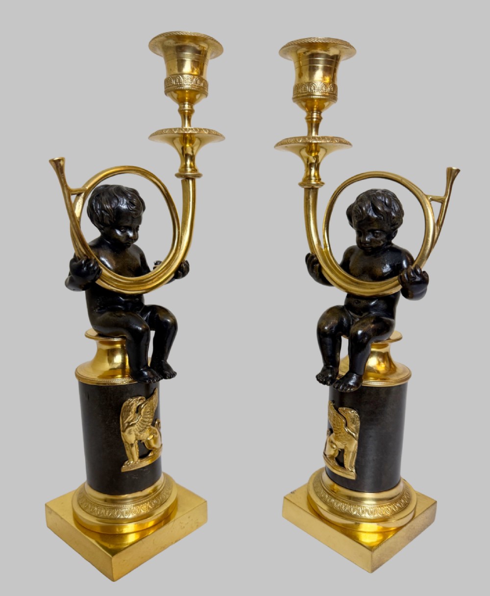 A pair of 19th-century gilt and patinated bronze candlesticks featuring dark cherubs seated on cylindrical pedestals with gilt winged lion motifs, holding gold-toned horn sconces. View VI