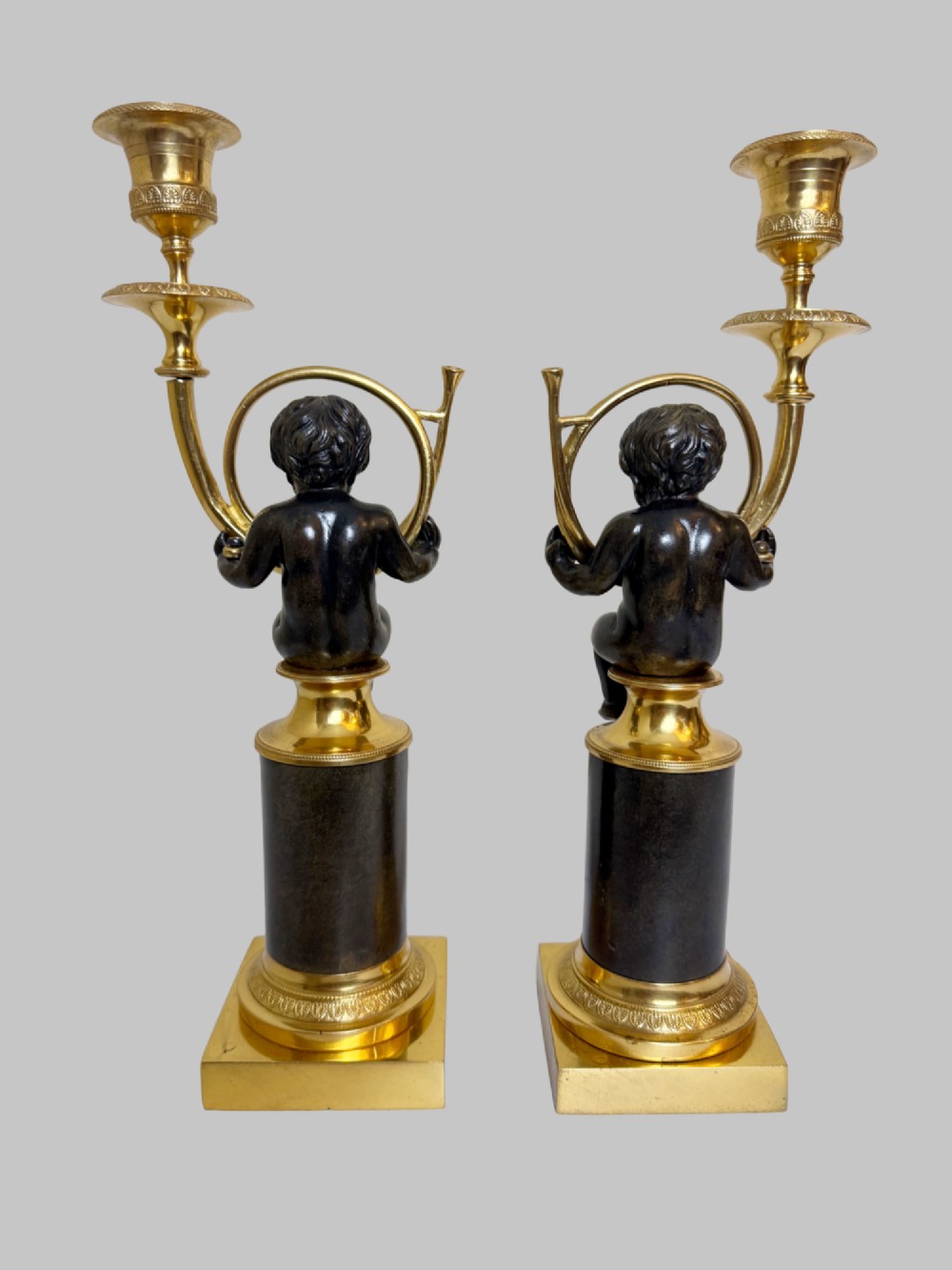 A pair of 19th-century gilt and patinated bronze candlesticks featuring dark cherubs seated on cylindrical pedestals with gilt winged lion motifs, holding gold-toned horn sconces. View IX