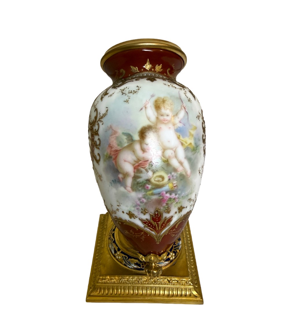 A 19th-century porcelain vase featuring hand-painted cherubs, deep red ground panels, gilded filigree, and a square gilt-bronze foot with enamel accents.