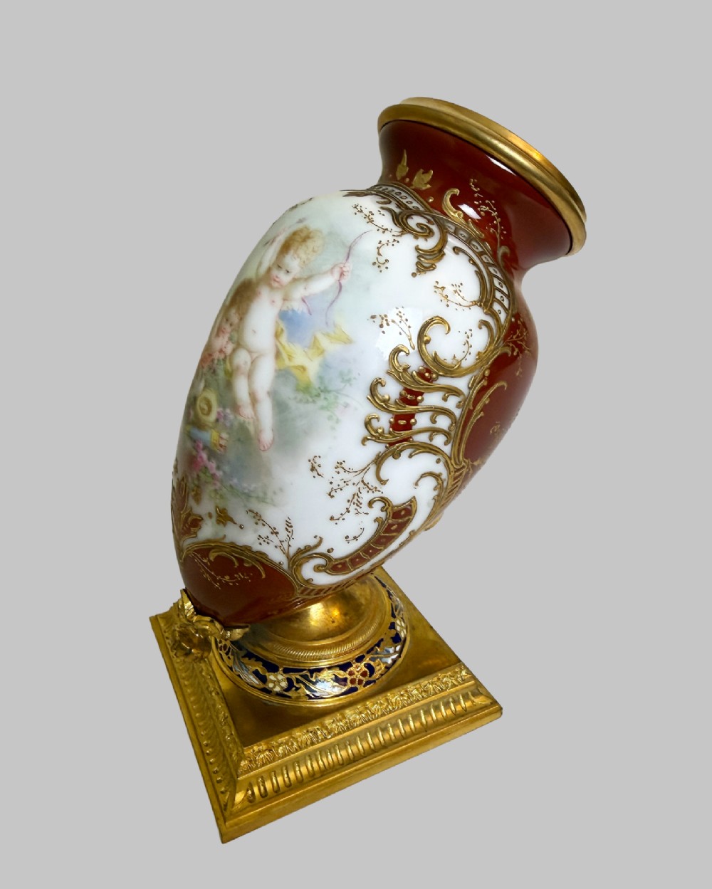 A 19th-century porcelain vase featuring hand-painted cherubs, deep red ground panels, gilded filigree, and a square gilt-bronze foot with enamel accents. View II