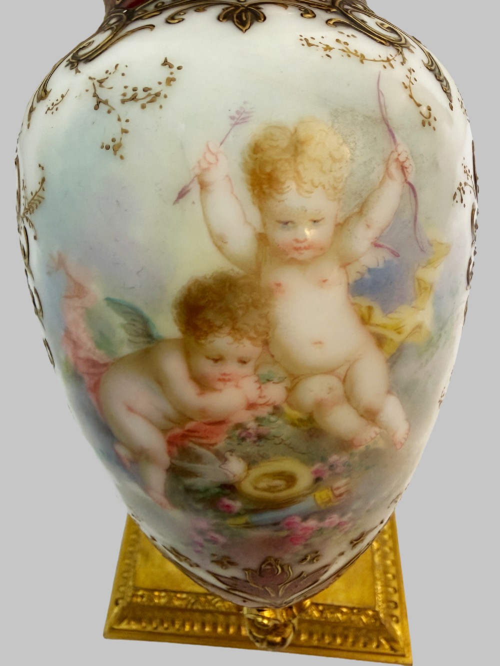A 19th-century porcelain vase featuring hand-painted cherubs, deep red ground panels, gilded filigree, and a square gilt-bronze foot with enamel accents. View V