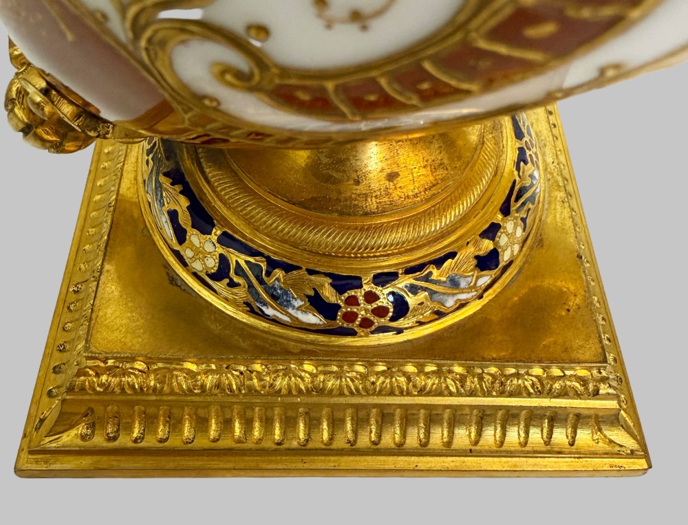A 19th-century porcelain vase featuring hand-painted cherubs, deep red ground panels, gilded filigree, and a square gilt-bronze foot with enamel accents. View VI