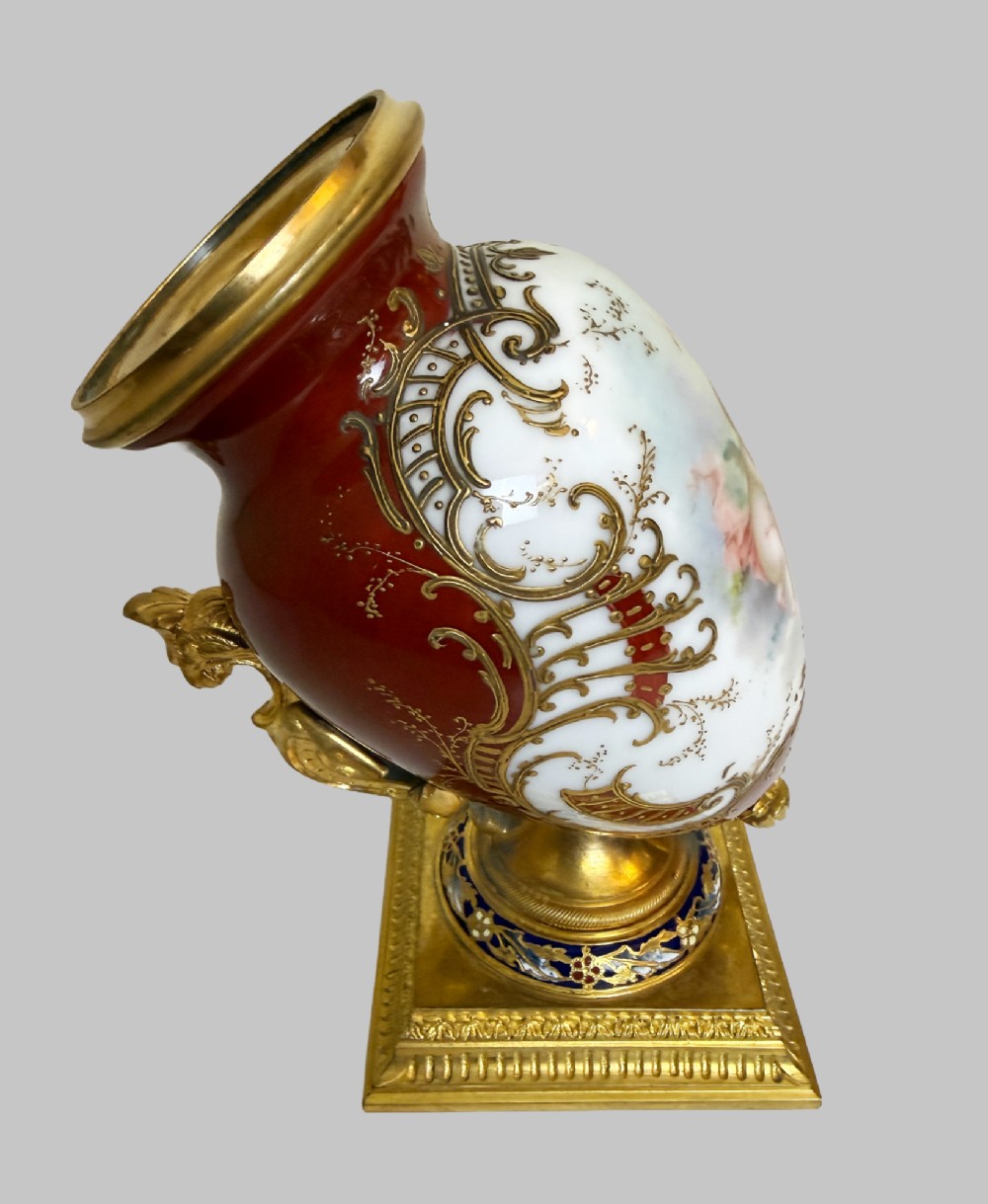 A 19th-century porcelain vase featuring hand-painted cherubs, deep red ground panels, gilded filigree, and a square gilt-bronze foot with enamel accents. View VII