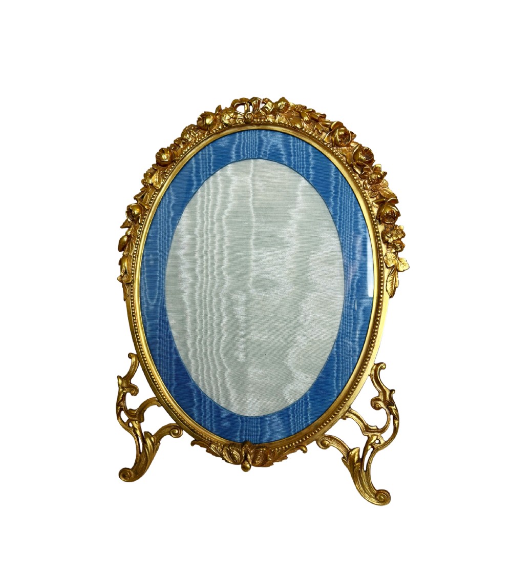 An antique oval gilt bronze picture frame featuring relief floral garlands, scrollwork legs, and a blue moire silk mount.