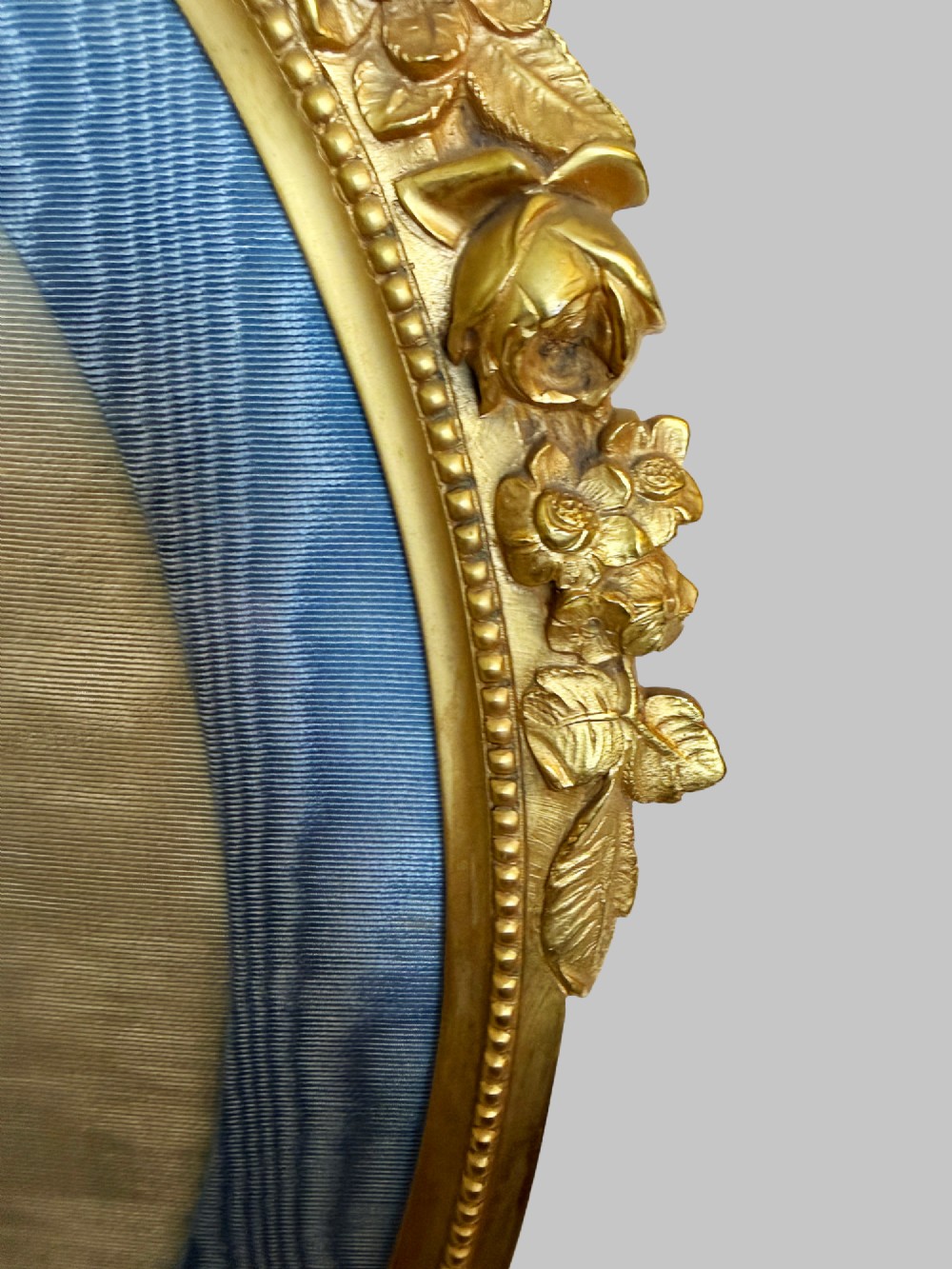 An antique oval gilt bronze picture frame featuring relief floral garlands, scrollwork legs, and a blue moire silk mount. View II