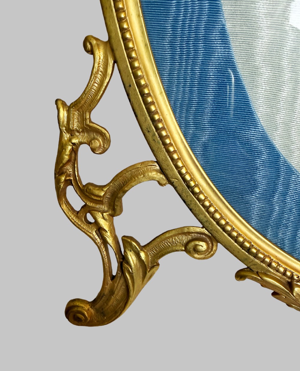An antique oval gilt bronze picture frame featuring relief floral garlands, scrollwork legs, and a blue moire silk mount. View III