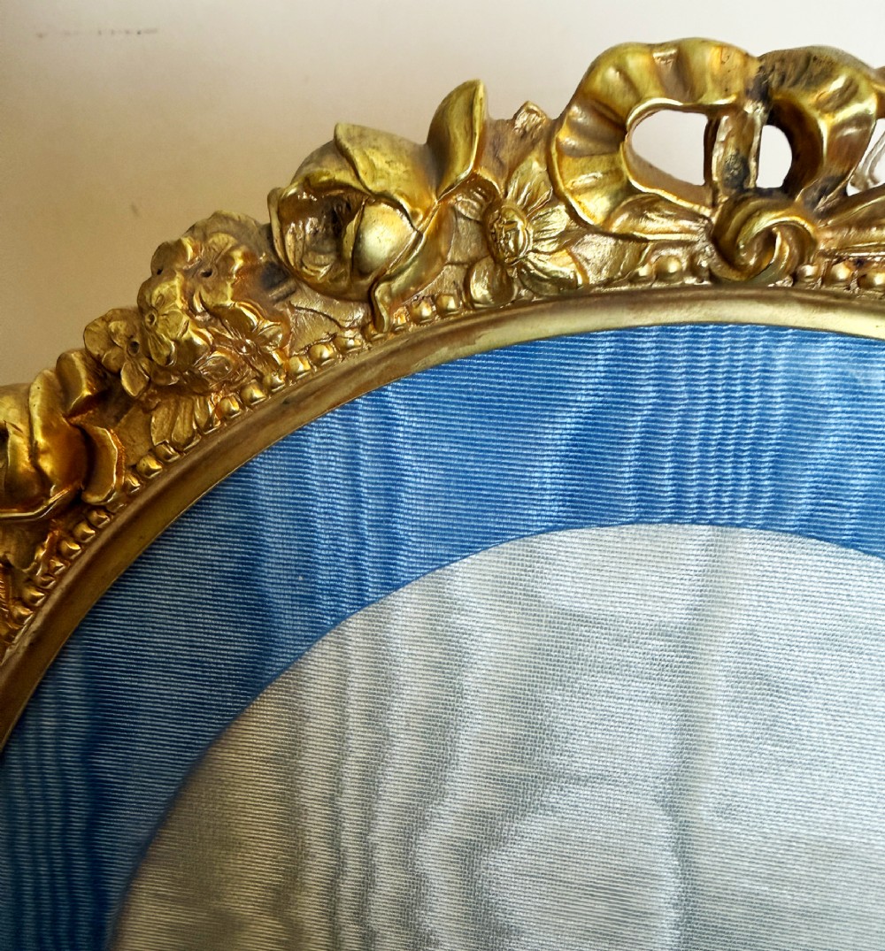 An antique oval gilt bronze picture frame featuring relief floral garlands, scrollwork legs, and a blue moire silk mount. View V