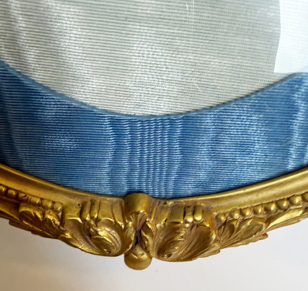 An antique oval gilt bronze picture frame featuring relief floral garlands, scrollwork legs, and a blue moire silk mount. View VI