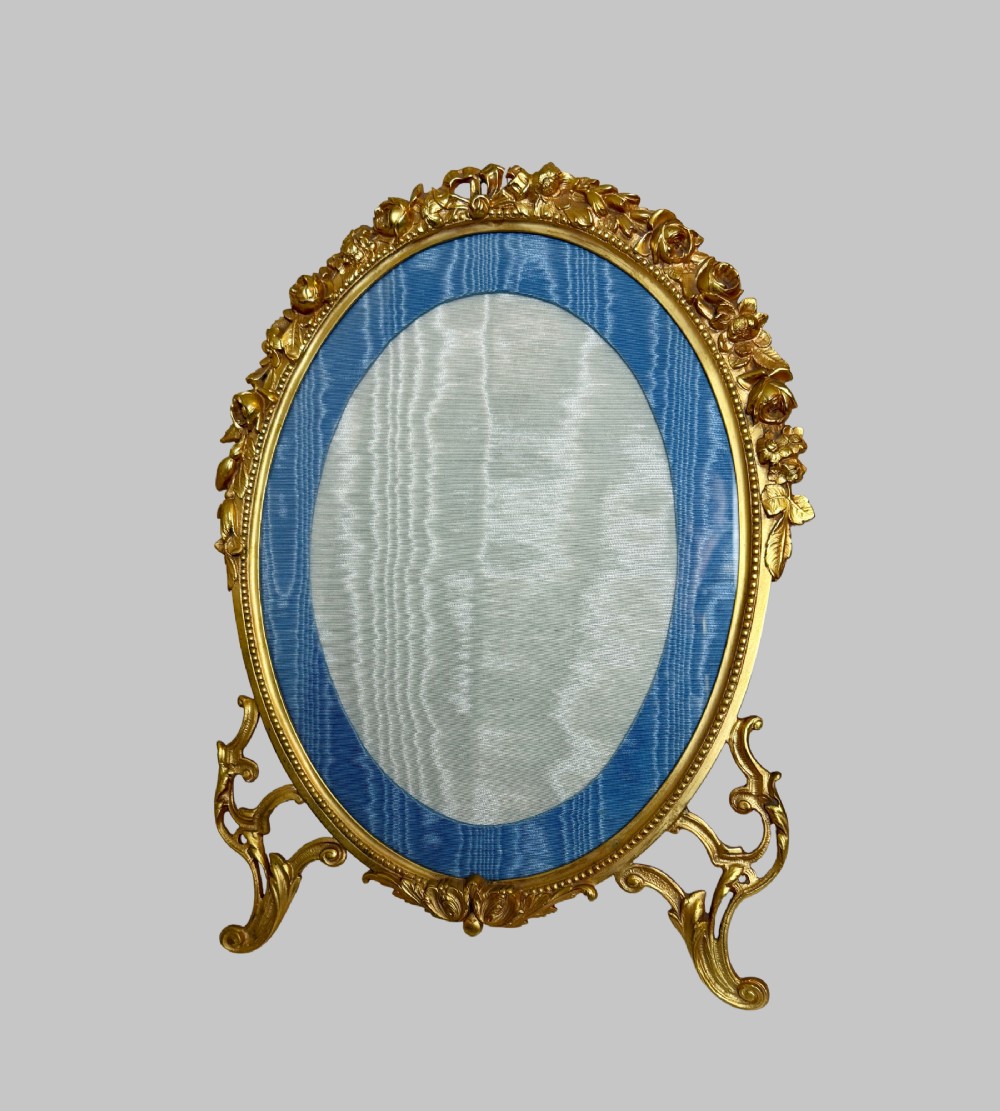 An antique oval gilt bronze picture frame featuring relief floral garlands, scrollwork legs, and a blue moire silk mount. View VII