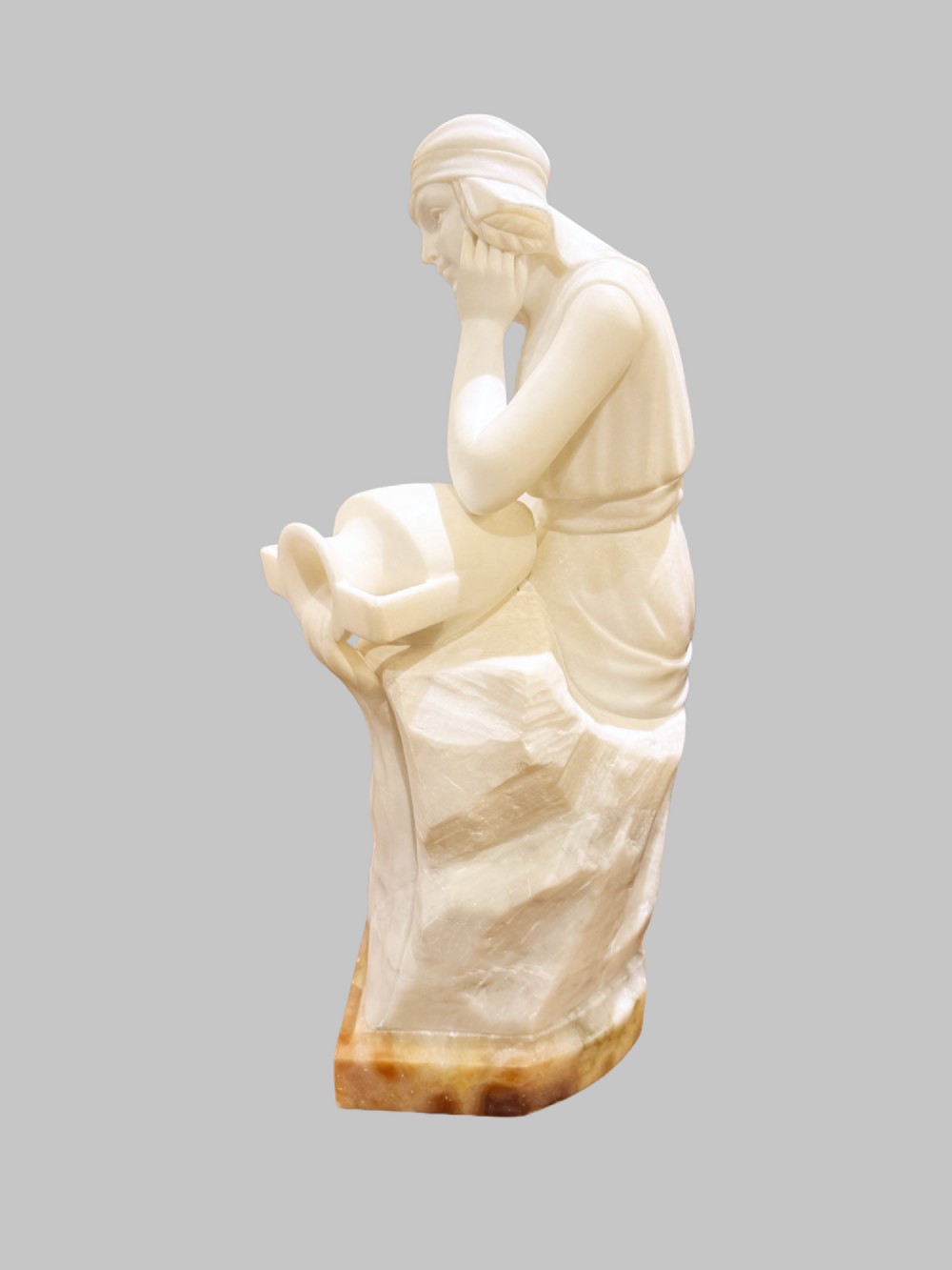 A carved white alabaster sculpture depicting a seated young maiden resting her chin on her hand beside a pitcher, mounted on a rectangular honey-coloured marble base. View VI