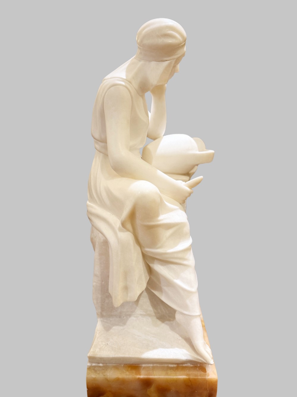 A carved white alabaster sculpture depicting a seated young maiden resting her chin on her hand beside a pitcher, mounted on a rectangular honey-coloured marble base. View VIII