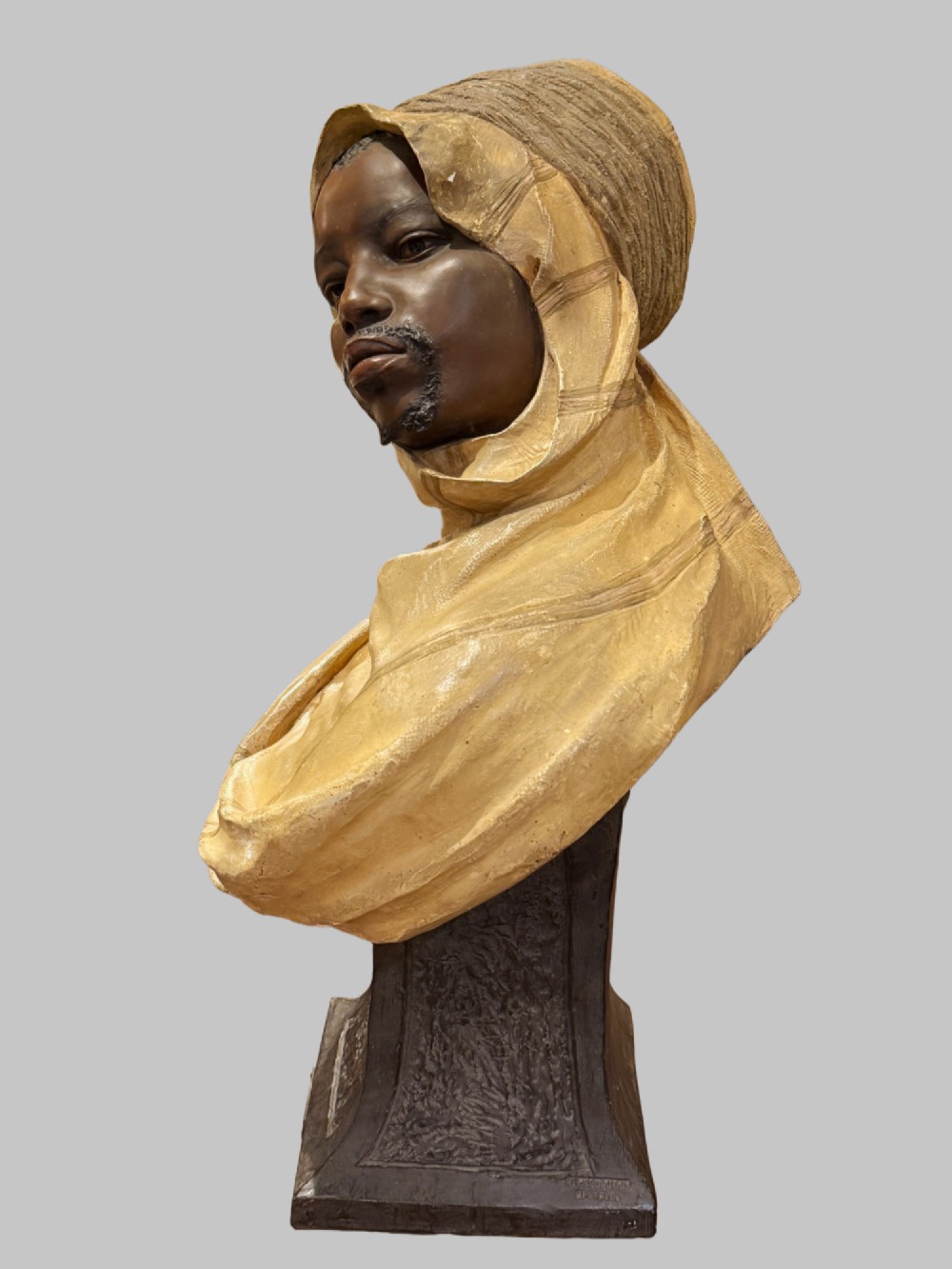 A cold-painted polychrome terracotta bust of a young North African man wearing a draped yellow headscarf mounted on a dark pedestal base. View X