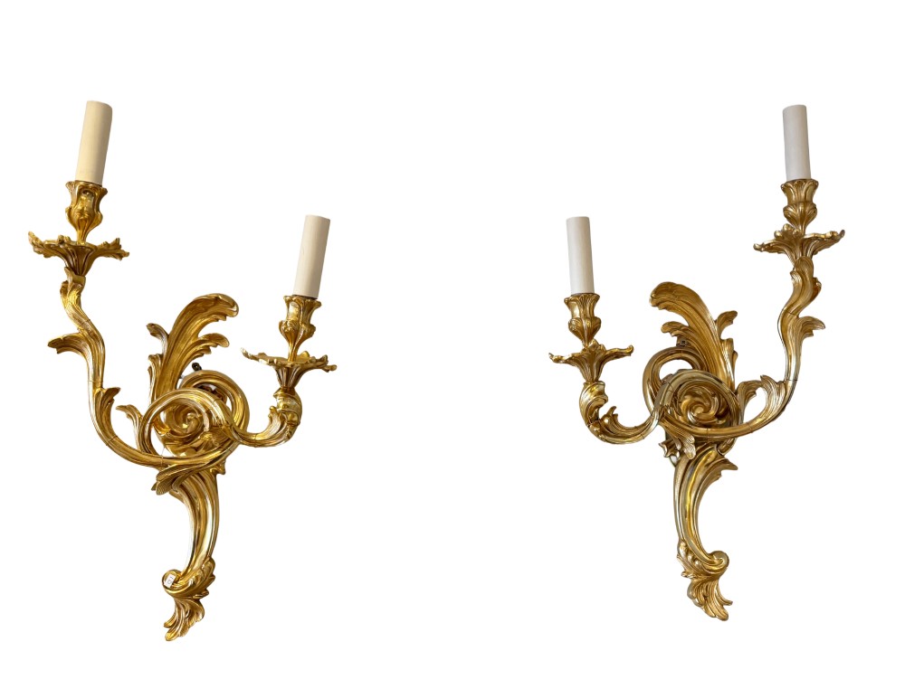 A pair of gilt bronze two-light wall sconces featuring elaborate Rococo scrolling acanthus leaf metalwork and cream candle sleeves.