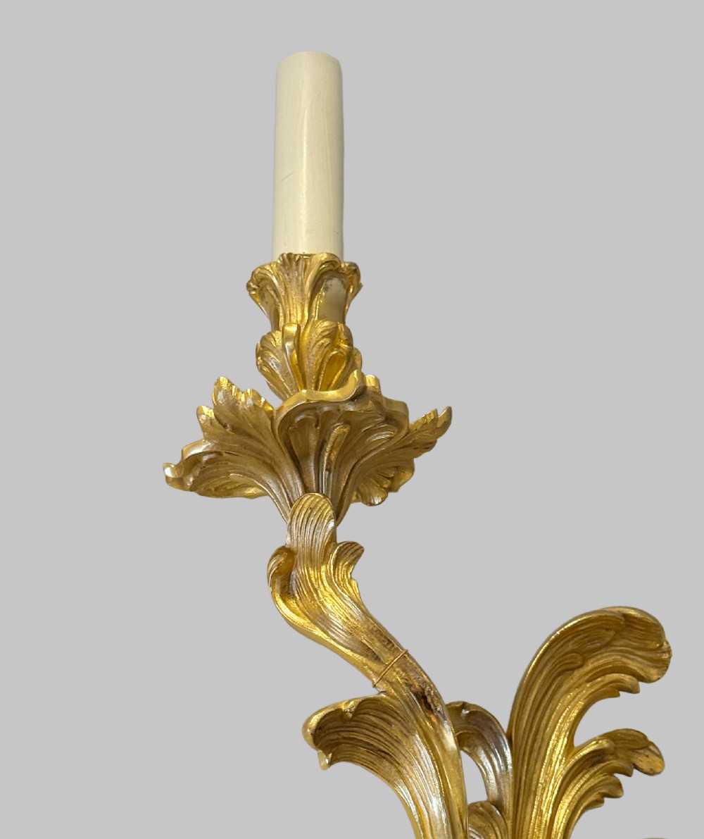 A pair of gilt bronze two-light wall sconces featuring elaborate Rococo scrolling acanthus leaf metalwork and cream candle sleeves. View IV