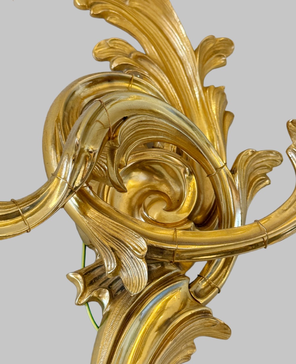 A pair of gilt bronze two-light wall sconces featuring elaborate Rococo scrolling acanthus leaf metalwork and cream candle sleeves. View V