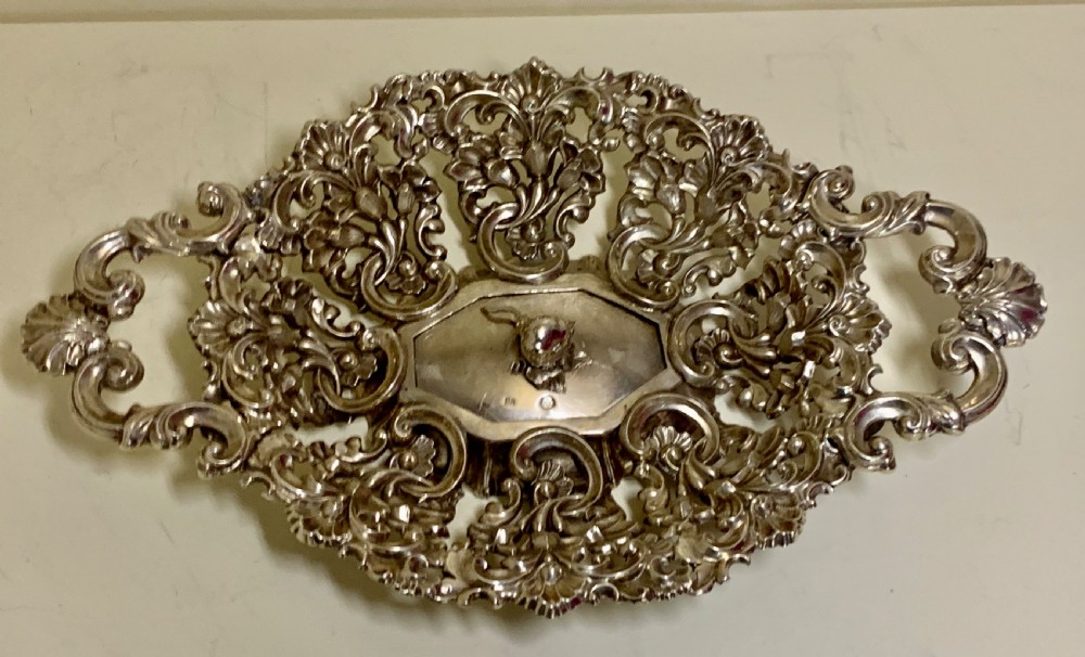 An oval antique silver basket featuring intricate openwork scrollwork, foliage motifs, and two integrated side handles.