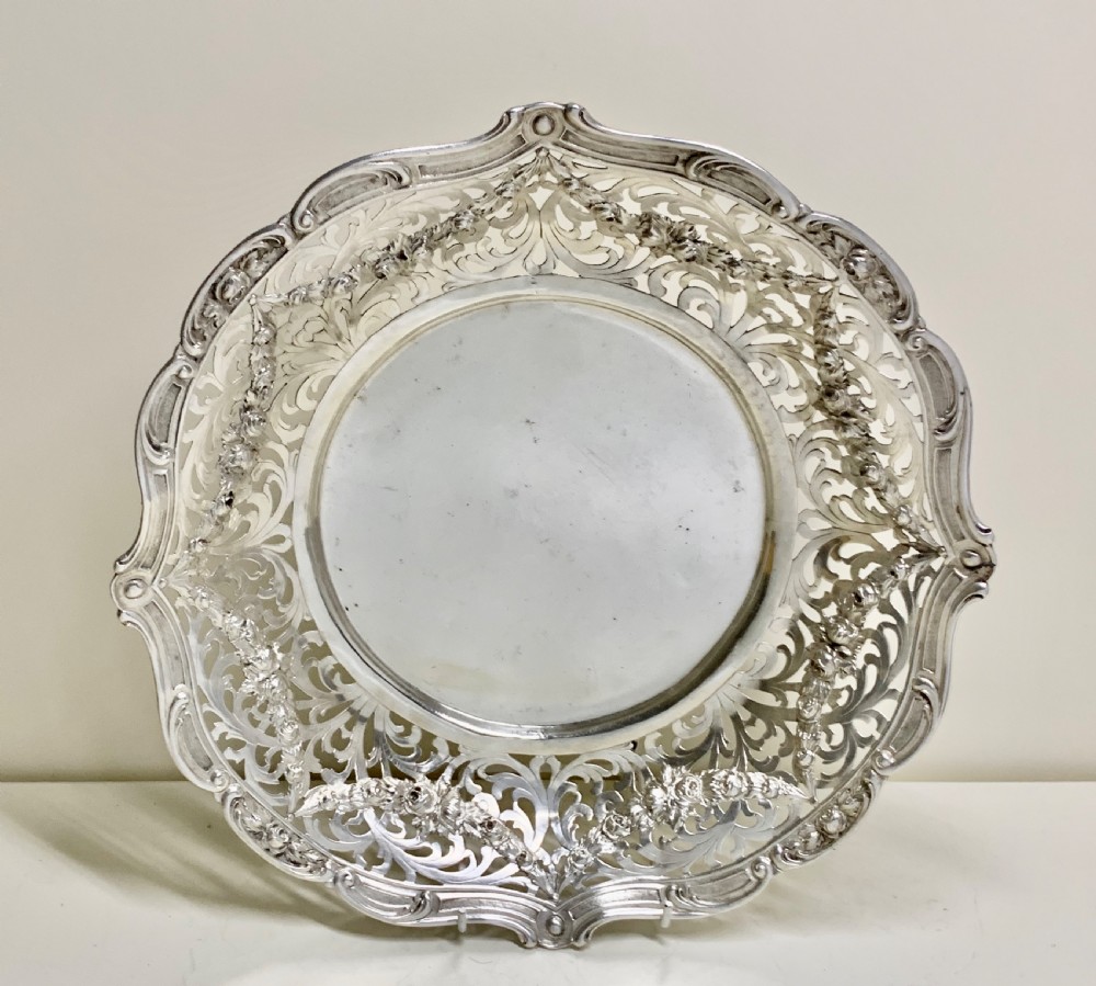 A circular antique solid silver bread basket featuring intricate hand-pierced fretwork, repousse floral swags, and a contoured scalloped rim.