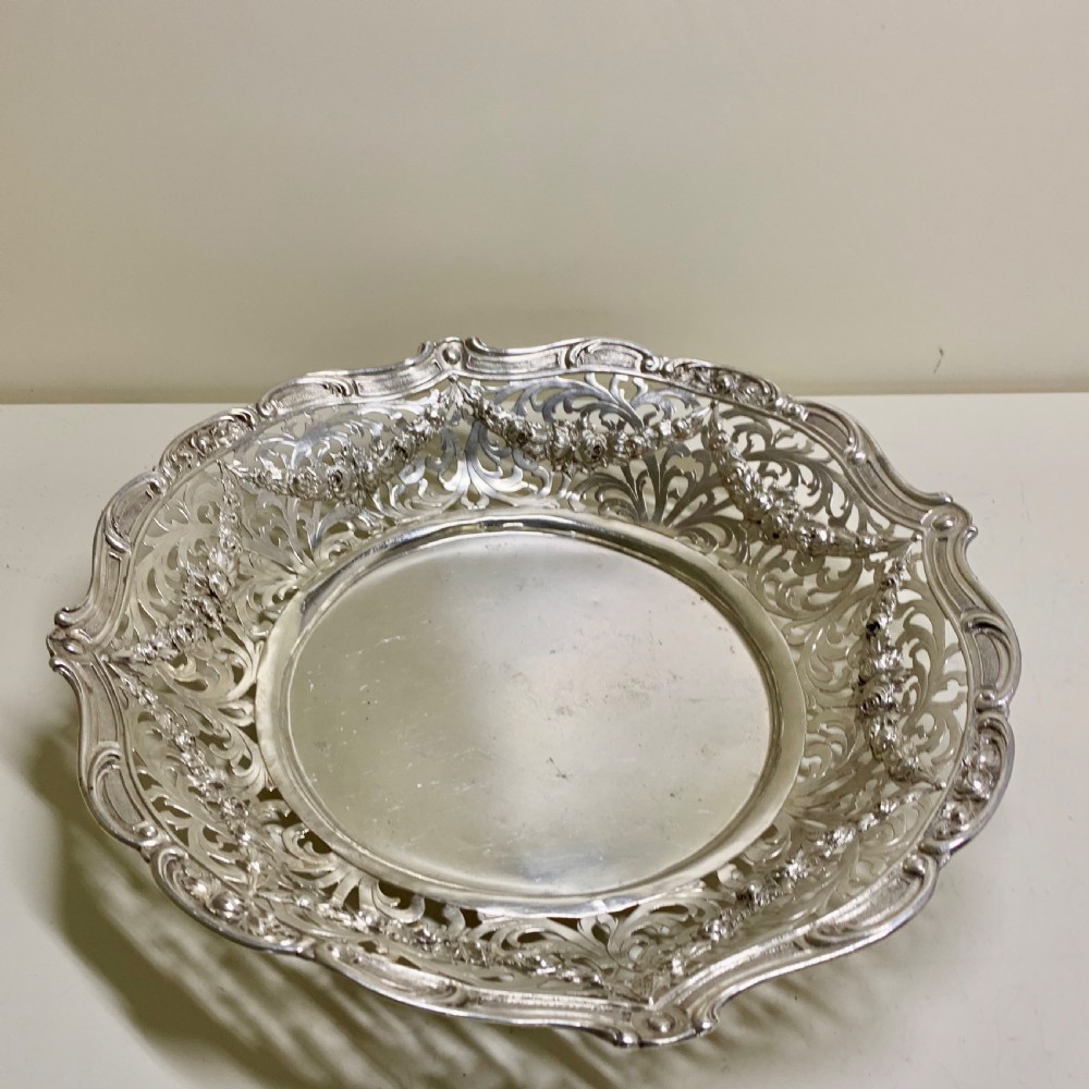 A circular antique solid silver bread basket featuring intricate hand-pierced fretwork, repousse floral swags, and a contoured scalloped rim. View IX