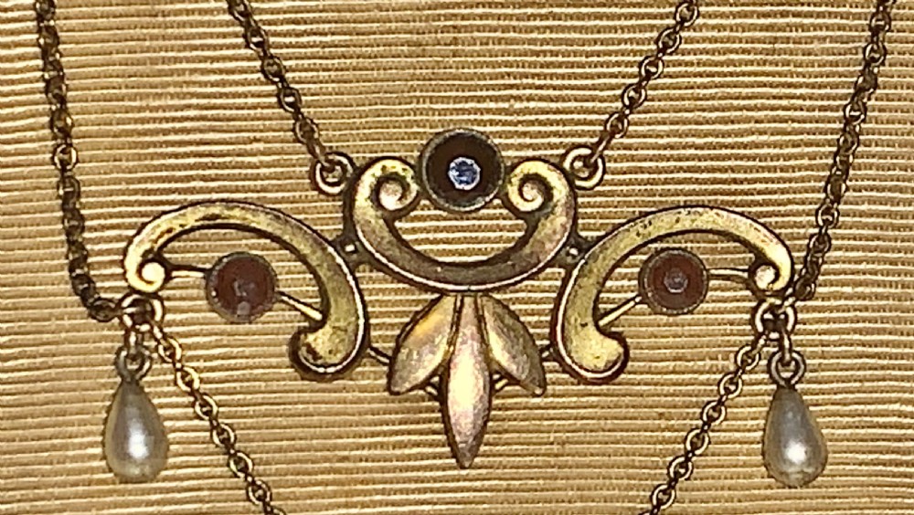 A Victorian 9ct gold lavaliere necklace featuring openwork scrolled motifs, central bezel-set round garnets, and hanging seed pearl drops. View IX